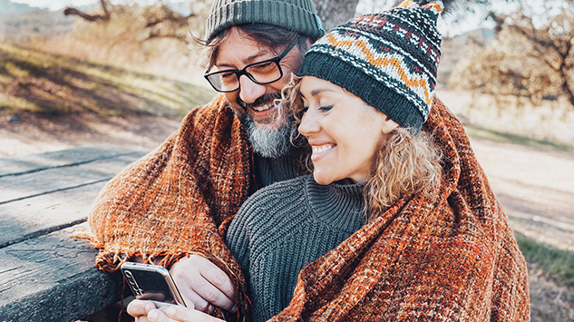 A couple wearing beanies outdoors sharing a throw blanket looking at their phone