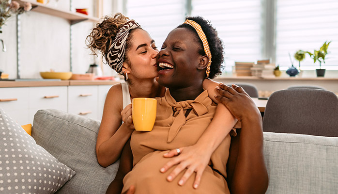A woman embracing her pregnant partner from behind while her partner is sitting on the couch
