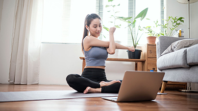 Woman sitting cross-legged on her yoga mat in front of her laptop while stretching her arm 