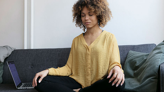 A woman sitting cross-legged on her couch with her hands on her knees meditating